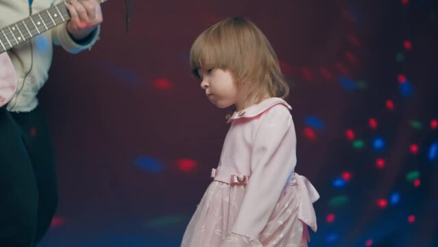 Little girl on stage in pink vintage dress, she dances, her father plays acoustic guitar. Color music is shining. Performance on stage. Early development of children