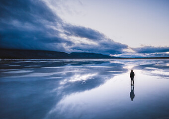 Lone figure stands on frozen lake with reflection and storm clouds