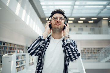 Fototapeta premium Student, library and man with music headphones in university, college or school. Thinking, education scholarship and face of young male streaming educational podcast, radio or song, audio or album.