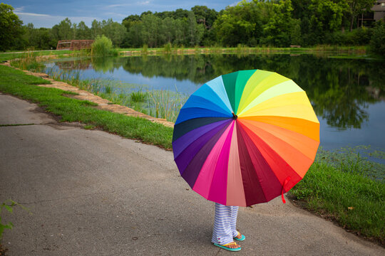 Faceless girl holding a rainbow umbrella on a path by a pond
