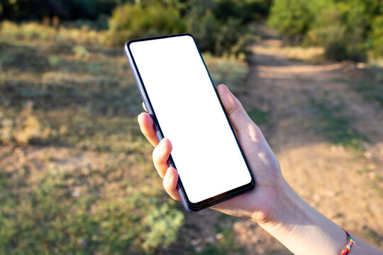 Hand holding phone with blank white screen while walking in the nature. Mockup image of smartphone.