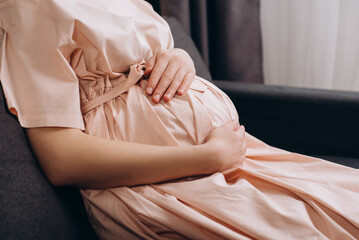 Close up of loving young pregnant woman sitting on couch stroking big belly, expressing love and care to unborn baby, expecting childbirth, feeling tenderness, resting alone at home. Maternity concept