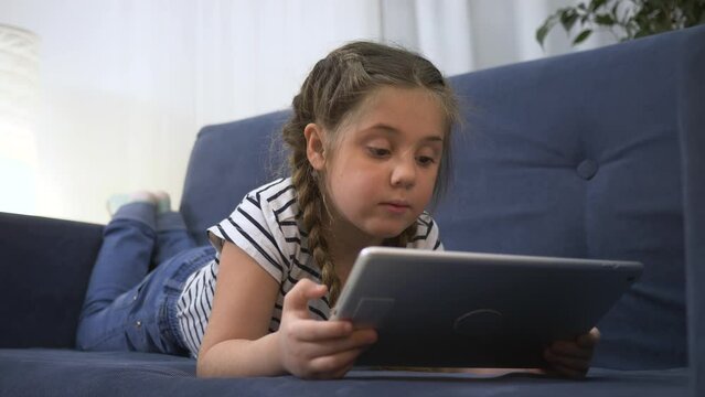 A lonely child loves to play on tablet while sitting on couch at home. The girl watches cartoons in everyday life, having fun from loneliness. child uses apps on tablet addiction concept