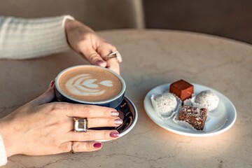 A cup of fresh cappuccino coffee in the hands of a woman on a fashionable background of a white marble table, next to a plate with sweets. Coffee addiction