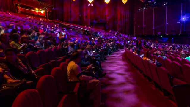 Spectators gather in the auditorium and watch the show in theatre timelapse. Large hall with red armchairs seats. Viewers filling places until turn off the light. Panoramic view from left side