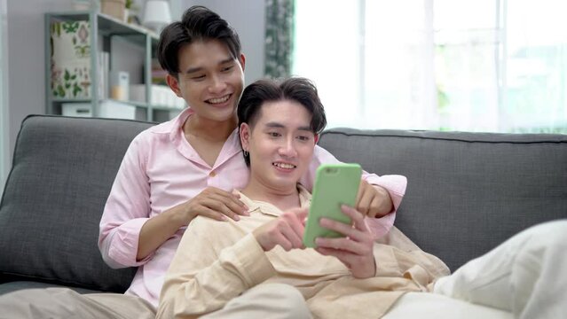 Cute Male Gay Couple Spend Time at Home. Young happy lgbtq couple Lying Down on a Sofa and Use a phone, doing online shopping in ecommerce app together, having fun checking social media together