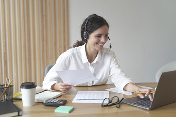 Happy woman wears headphones while sits at desk at home office looks at laptop screen works online