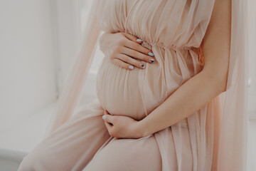 Cropped image of faceless woman touches her abdomen anticipates for baby wears festive dress poses indoor. Women and pregnancy concept