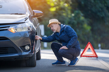 elderly man Sitting on the side of the road near a broken car and checking, the tire for a problem.