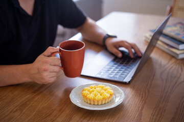 A white man wearing a black t-shirt working in his desk at home using a personal computer and holding a cup of coffee next to a delicious lemon pie during the day