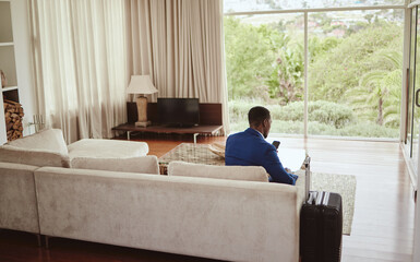 Phone, travel and hospitality with a business black man sitting on a sofa in the living room of a...