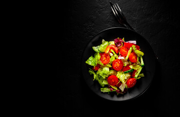 various fresh mix salad leaves with tomato, cucumber and red pepper on a black plate on Dark grey black slate background