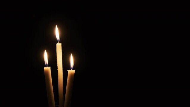 Three candles are lit on a black background, close-up. The yellow flickering flames illuminate the darkness. The warm glow of flame burning at night and moving with the wind. Isolated, Copyscape. 4K