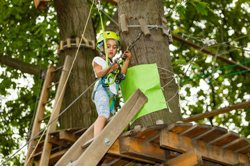 Portrait of little smiling girl in helmet and harness on trail in sky rope park in summer.