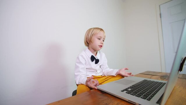 Caucasian blue-eyed boy in stylish black tie bow and bright trousers looks at the laptop screen and talks to someone. Online speech lesson at home for a 5 years old boy