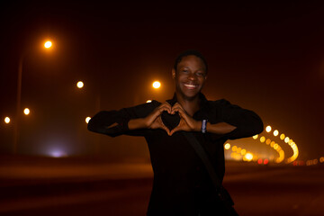 smiling young african man on a black t-shirt showing shaped heart with his hands, looking at the camera at nigh on the street