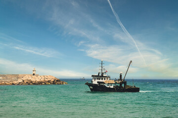 Obraz premium View of a fishing vessel returning from the sea to land and a view of the small harbor, lighthouse and gorgeous dramatic sky. 