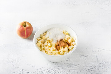 White ceramic bowl with sliced apples with sugar and cinnamon for apple pie filling on a light gray background