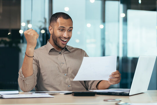 Portrait of successful hispanic businessman, man in shirt reading report and happy celebrating success satisfied with result holding hand up, worker inside office with laptop working on paper work.