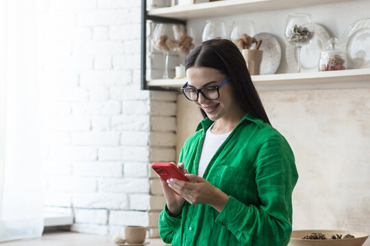 A young beautiful woman is standing at home in the kitchen near the window with a phone in her hands. orders food home, makes purchases in an online store.