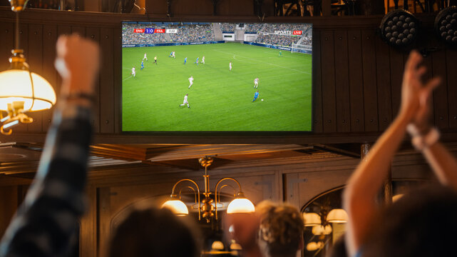 Group of Soccer Fans Watching a Live Football Match Broadcast in a Sports Pub on TV. People Cheering, Supporting Their Team. Crowd Goes Ecstatic When Team Scores a Goal and Wins the Championship. - Powered by Adobe