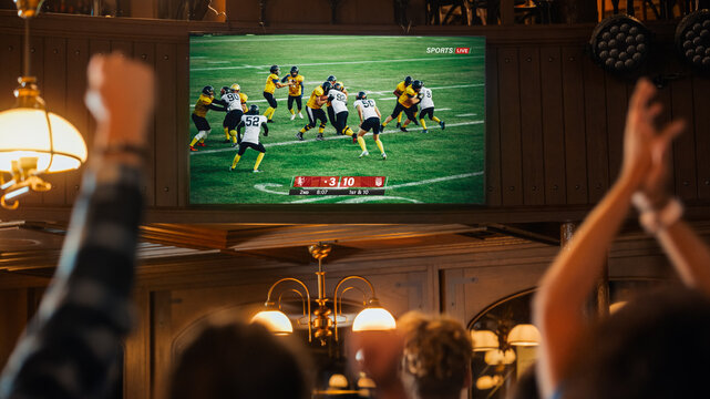 Group of American Football Fans Watching a Live Match Broadcast in a Sports Pub on TV. People Cheering, Supporting Their Team. Crowd Goes Ecstatic When Team Scores a Goal and Wins the Championship.