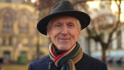 Close-up grey-haired senior man putting on elegant hat looking at camera smiling. Portrait of handsome confident Caucasian male retiree posing on city street on sunny spring autumn day