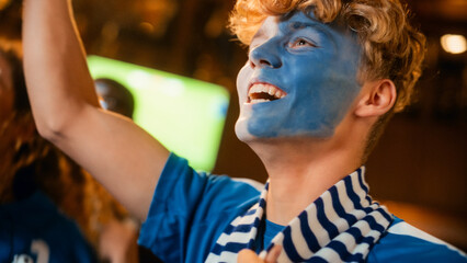 Close Up Portrait of a Handsome Young Soccer Fan with Painted Blue and White Face Standing in a Crowd in a Bar, Chanting, Jumping, Cheering for a Football Team. Friends Celebrate the Goal. Slow Motion