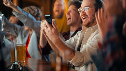 Portrait of an Excited Young Man Sitting at Bar Counter, Drinking Beer and Using a Smartphone, Anxious About a Sports Bet on His Favorite Soccer Team. Joyful Emotions When Football Team Scores a Goal.