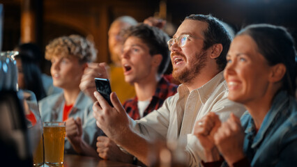 Portrait of a Happy Young Man Holding a Smartphone, Anxious About the Sports Bet on a Soccer Match. Looking at the Screen While Sitting at a Counter, Getting Very Emotional After Winning the Bet.