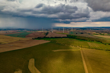 Drone flight at high altitude over a large field with many wind turbines spinning. The wind turbines cast a long shadow because the sun is setting. In the background, at storm sky.