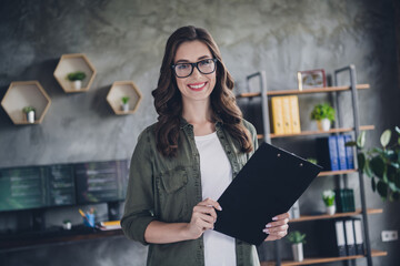 Photo of smiling positive lady hacker wear spectacles holding clipboard list indoors workplace workshop
