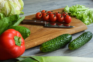 Closeup fresh organic vegetables with kitchen knife on cutting board wooden table indoors. Composition of red green food ingredients for cooking healthy vegetarian eating at home. Preparing meal salad
