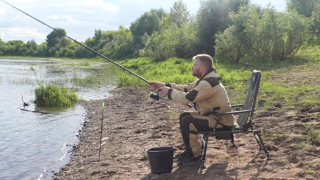 Side view of young bearded fisherman wearing raincoat sitting on river bank on travel chair with fishing rod catches fish, slow motion. Calm and peaceful picture. Lifestyle, leisure activity in nature