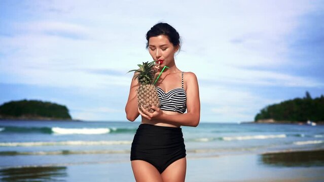 Female tanned asian woman drinking. beautiful woman in black bikini with pineapple in hands on beach tropical background with copyspace. healthy eating, fructorianism and vegetarianism, summer vitamin