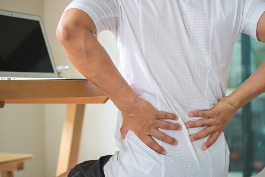 Working Man Sitting On The Desk In The Living Room At Home. He Used Both Hands Holding To Press Down On The Lower Back. He Is Suffering From Back Pain From Sitting For A Long Time.