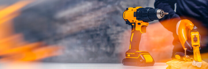 Man worker holds an electric screwdriver in his hands close-up against the background of a construction tool and a concrete wall. Long banner with glow effects.