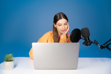 Teen podcaster looking at laptop near microphone isolated on blue.