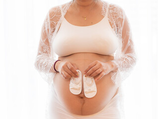 Closeup pregnant belly with cute tiny baby shoes. Concept of pregnancy, gynecology, preparation. Beautiful pregnant woman in shirt in white background. Woman holding big pregnant stomach.
