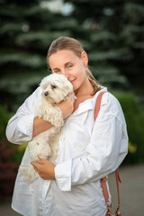 Portrait of a young beautiful blonde with a Maltese in her arms on a summer day outdoors.