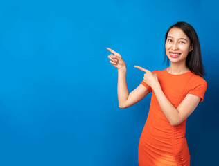 Asian Happy beautiful slim smiling Chinese woman pointing side. Positive relaxed  mood. Orange dress and blue background. horizontal banner copyspace