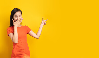 Silly laughing beautiful Asian woman in bright orange dress in yellow background looking in camera, covering hiding her face pointing finger side. Making jokes. Long horizontal banner copyspace