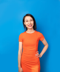 Asian Chinese beautiful woman in orange dress on blue background. Slim happy girl standing side  and looking at camera. 