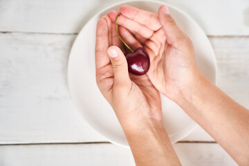 A close-up of a ripe large cherry in the shape of a heart in the hands of a girl, against a white plate. The concept of healthy nutrition and vitamins with space to copy. High quality photo