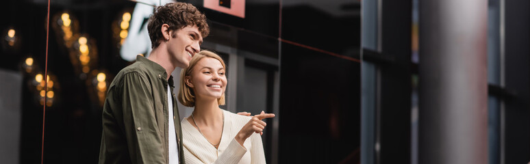 Fototapeta premium joyful blonde woman looking away and pointing with finger near boyfriend in hotel hall, banner.