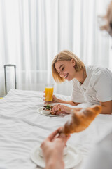 cheerful blonde woman having breakfast with orange juice and croissant on hotel bed near blurred boyfriend.