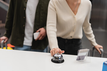 partial view of woman ringing service bell on reception near blurred boyfriend with smartphone in hotel.