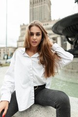 Elegant beautiful stylish woman model in fashion casual business clothes with a white shirt and black jeans sits near a vintage fountain in the city and looks at the camera