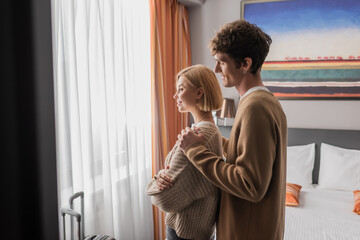 side view of smiling man hugging girlfriend standing with crossed arms near window in hotel room.