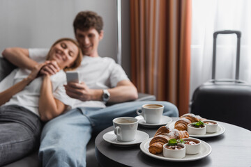 coffee cups and croissants with jam and chocolate paste near blurred couple with smartphone in hotel room.